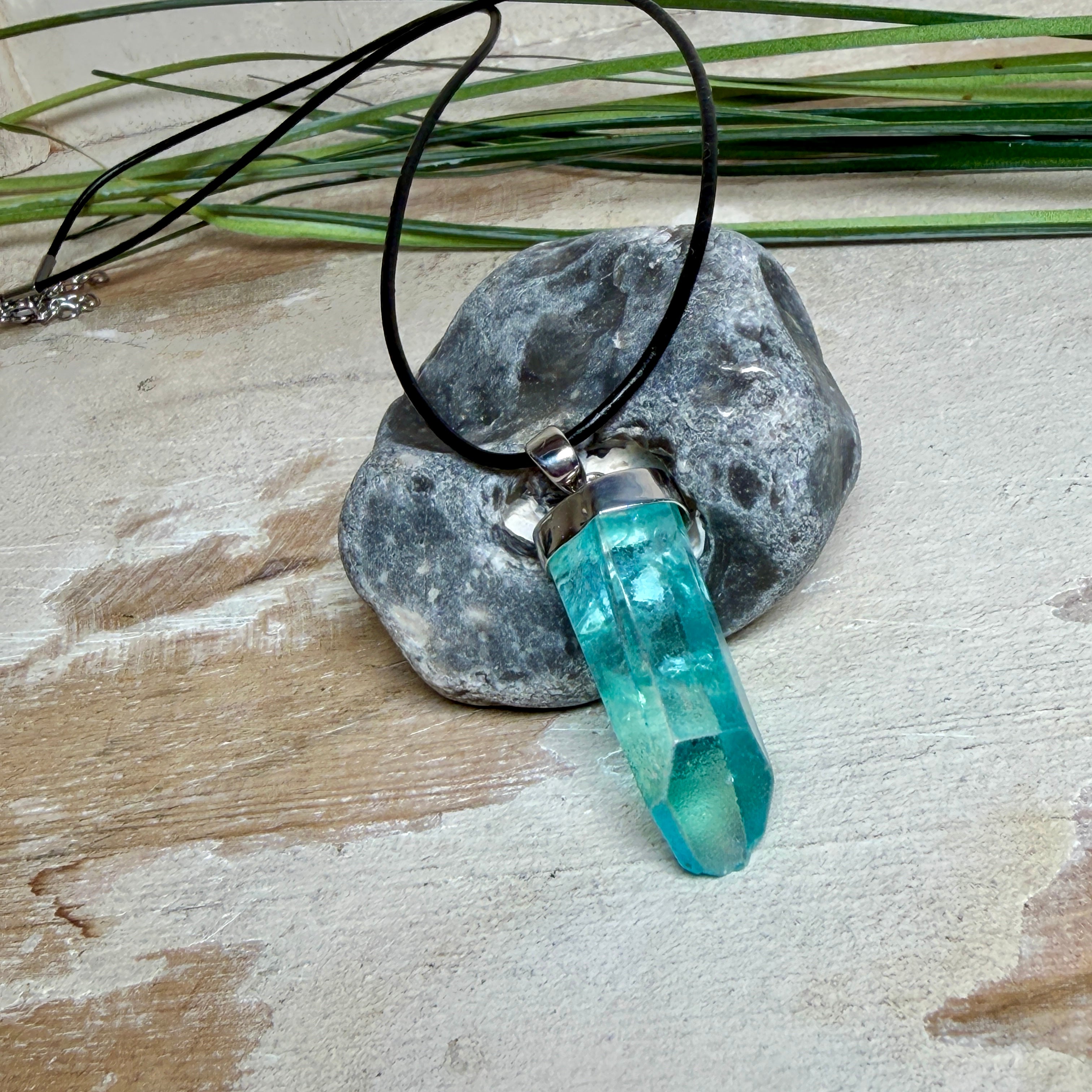 Nahaufnahme einer handgefertigten Halskette mit Aqua Aura Rohkristall: naturbelassener, sechskantiger Bergkristall mit goldbedampfter Aqua-Beschichtung in Türkisblau, gefasst in 925er Silber und getragen an einer schwarzen Lederkette mit Edelstahl-Karabiner und Verlängerungskettchen.