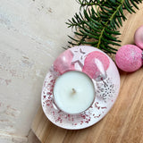 Rosa Teelichthalter aus klarem Epoxidharz, ca. 8 × 6,5 cm, Höhe ca. 4 cm. Im Harz sind kleine rosa Weihnachtskugeln, ein Stern, eine Schneeflocke und rosa Granulat eingegossen. Ovaler Halter für ein Teelicht, Unterseite mit vier runden Korkpads als Möbelschoner. Fotografiert auf Holz mit Tannenzweig und rosa Kugeln als weihnachtlicher Dekohintergrund.