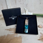 Rechteckiger Stabanhänger aus Maserholz und glasklarem Epoxidharz in tiefen Blau- und Türkistönen mit organischer Lagunen/Riff-Optik. Hochglanzpoliert, ca. 5,3 × 1,2 cm. Der Anhänger hängt an einer schwarzen Lederkette mit Edelstahl-Karabiner und 5-cm-Verlängerung (45 + 5 cm); die Anhängerschlaufe ist ebenfalls aus Edelstahl. Handgefertigtes Unikat, Lieferung in edler Schmuckbox – maritimer Look.