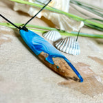 Handgefertigter Surfbrett-Anhänger „Ocean Glide“ aus Maserholz und Epoxidharz in verschiedenen Blau-, und Weißtönen. Der ca. 6,5 × 2 cm große Anhänger wird an einem verstellbaren Lederband (26–46 cm) mit Holzperlen geliefert. Ein maritimes Unikat mit natürlicher Holzmaserung, das die Schönheit des Meeres einfängt.
