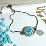 Verstellbares Edelstahlarmband mit Zirkoniasteinchen und Schiebeverschluss und rundem Anhänger (ca. 2,5 × 1,7 cm) mit echten Larimar-Edelstein-Splittern in Harz. Zusätzlich ein Charm „Blume des Lebens“ aus 925 Silber. Handgefertigtes Unikat in Türkis- und Meeresblautönen – Schmuckstück in Nahaufnahme bzw. als Tragebild am Handgelenk.