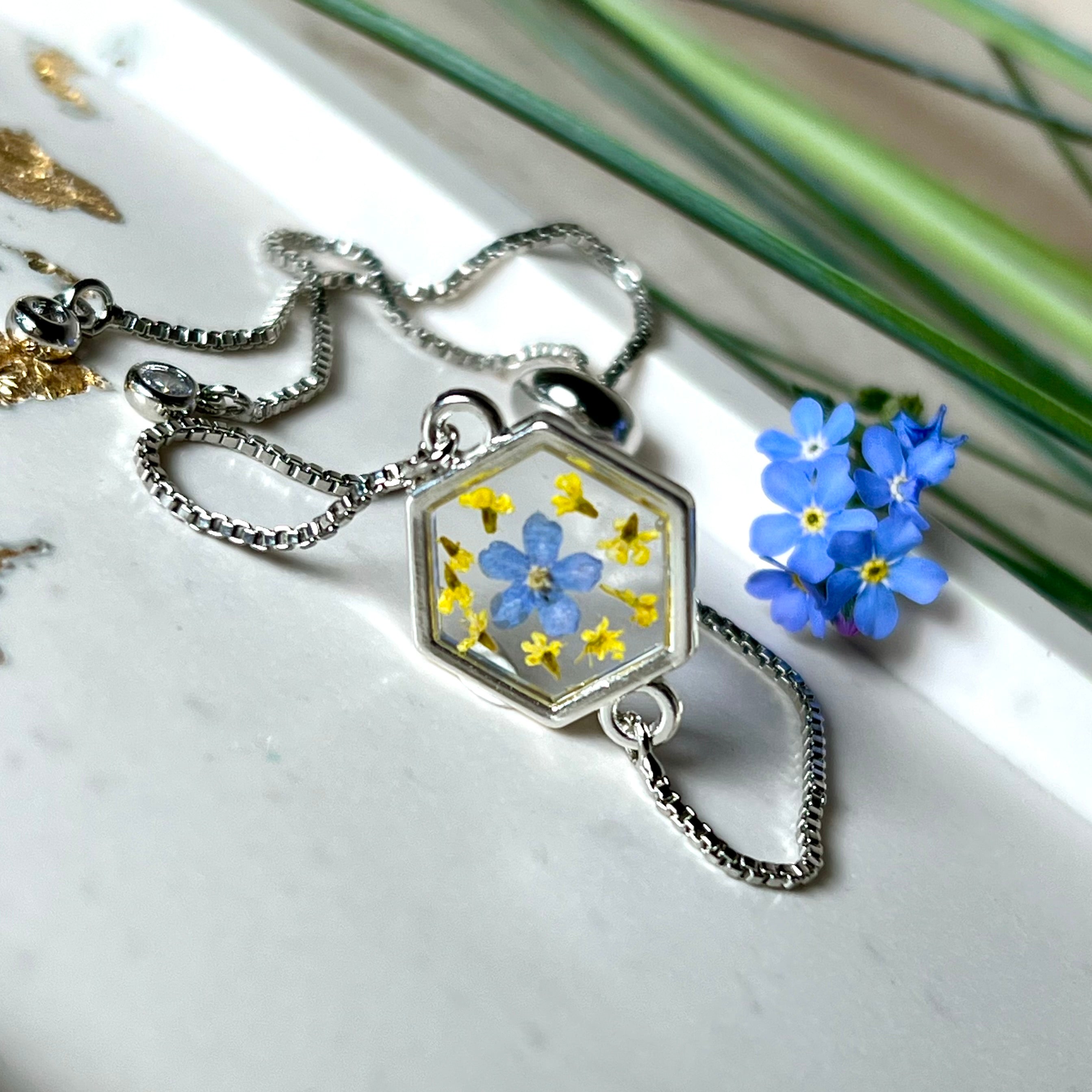 „Detailaufnahme eines handgefertigten Armbands mit echter blauer Vergissmeinnicht und feinen gelben Blüten in Harz, eingefasst in sechseckiger versilberter Lünette. Verstellbares Edelstahlarmband mit funkelnden Zirkonia-Endperlen – liebevolles Natur-Schmuckstück, ideal als Geschenk für Frauen.“