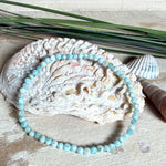 Zartes Larimar-Armband „Lagunenlicht“ mit facettierten 3 mm Perlen aus seltenem Atlantisstein auf Elastikfaden. Die kleinen Larimarsteine zeigen sanfte Blau- und Weißtöne wie Lagunenwasser. Innenumfang ca. 19 cm. Fotografiert auf Muscheln, einem dekorativen Stein und in einer schwarzen Schmuckbox von ZeitlosSchmuckDesign im maritimen Stil.
