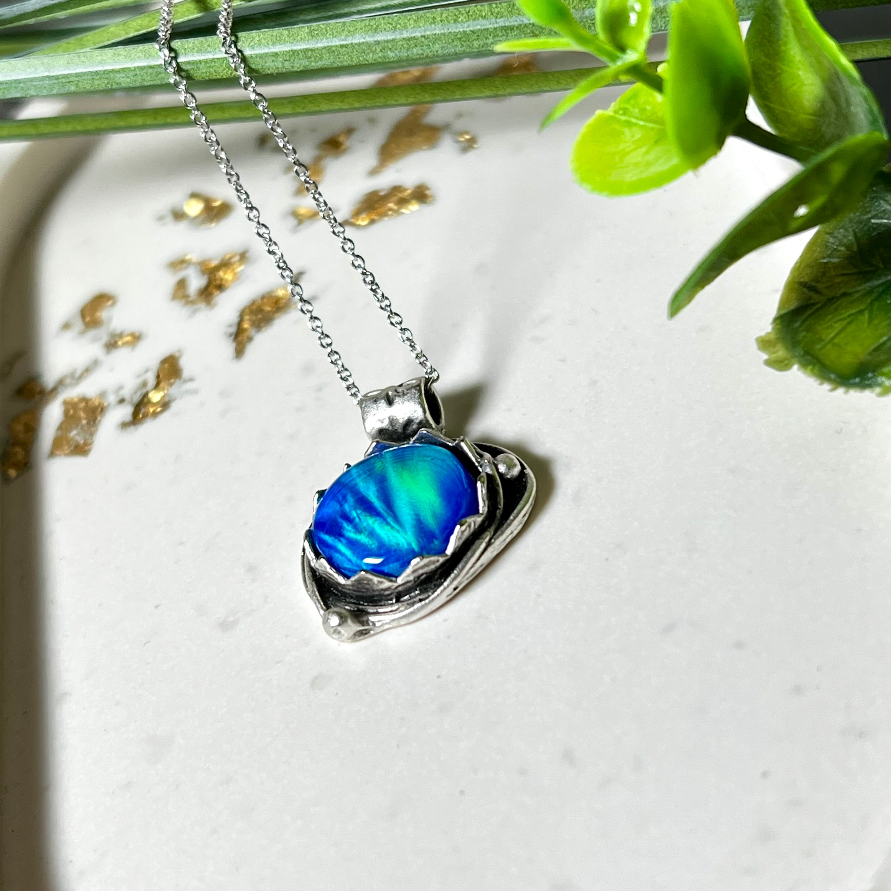 Handgemachter Anhänger mit rundem Cabochon aus Epoxidharz und schimmerndem synthetischem Opal. Die Farben changieren je nach Lichteinfall in Blau, Türkis und Grün. Der Opal sitzt in einer antik versilberten Fassung aus Messing (blei- & nickelfrei). Geliefert mit einer rhodinierten 925 Silberkette (45 cm). Jedes Stück ein Einzelstück – perfekt als stilvolles Geschenk oder persönlicher Glücksbringer.