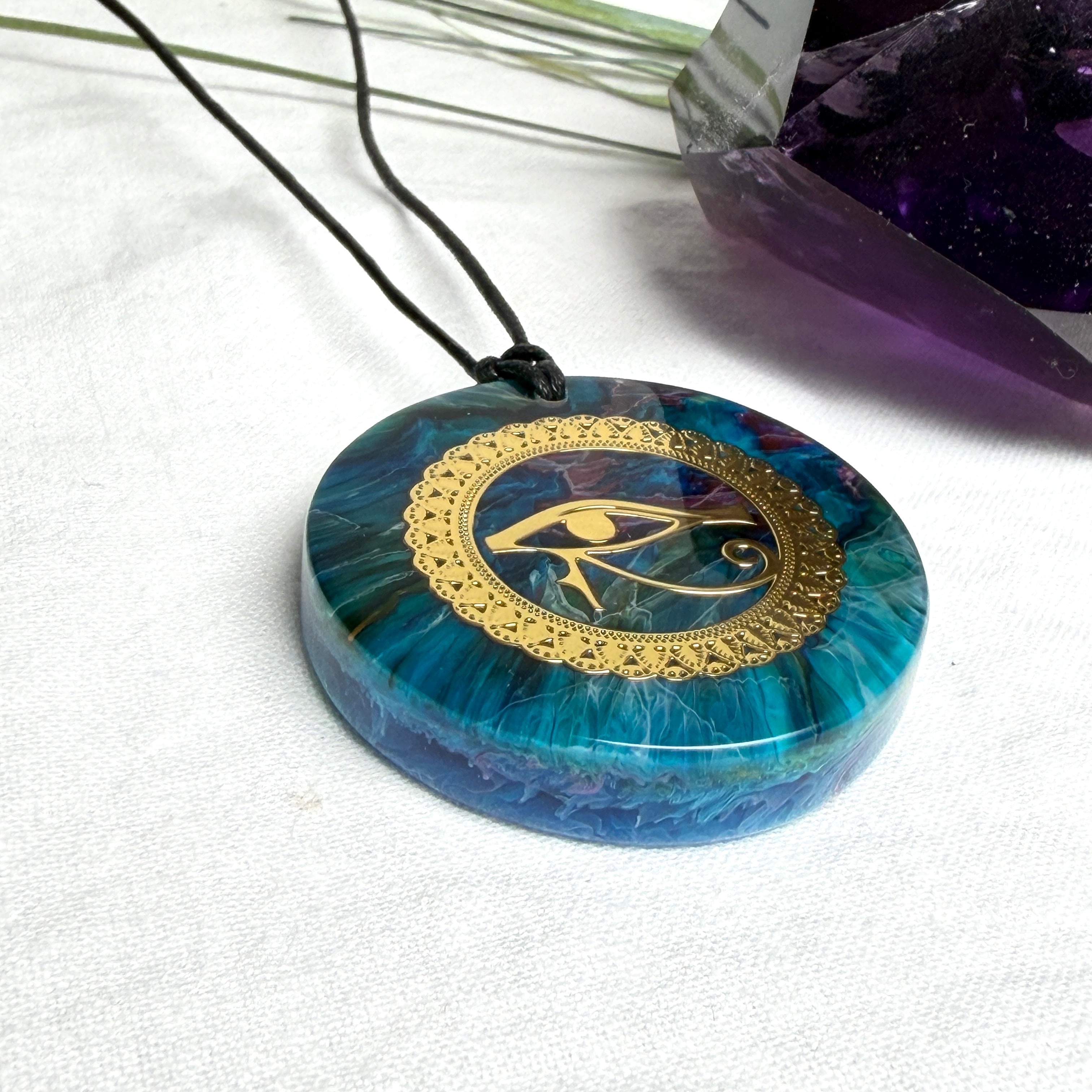 Runder Anhänger mit goldenem Horusauge aus Metall in blau türkis violettem Epoxidharz am schwarzen Baumwollband, spiritueller Eye of Ra Naturschmuck Amulett
