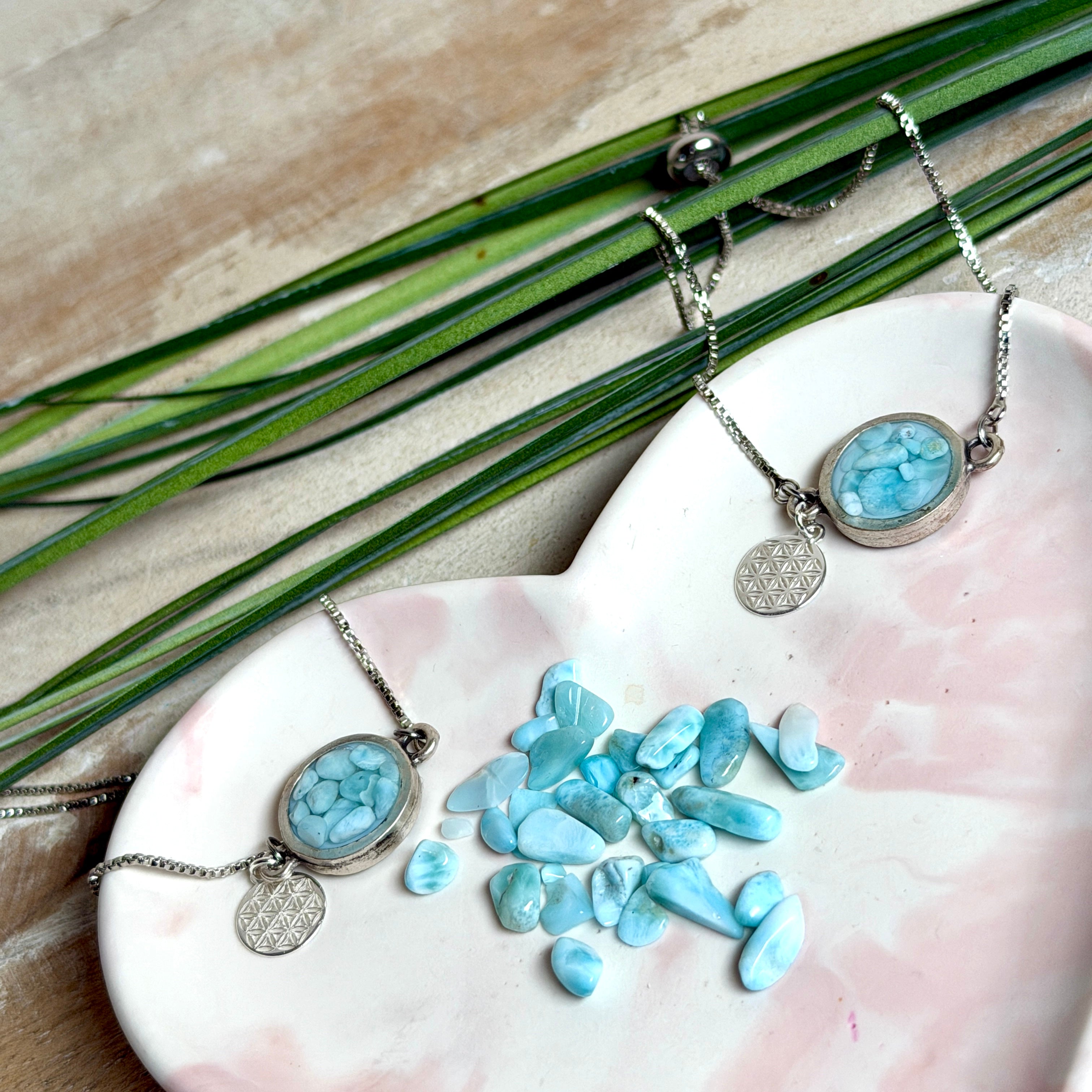 Verstellbares Edelstahlarmband mit Zirkoniasteinchen und Schiebeverschluss und rundem Anhänger (ca. 2,5 × 1,7 cm) mit echten Larimar-Edelstein-Splittern in Harz. Zusätzlich ein Charm „Blume des Lebens“ aus 925 Silber. Handgefertigtes Unikat in Türkis- und Meeresblautönen – Schmuckstück in Nahaufnahme bzw. als Tragebild am Handgelenk.