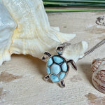 Feine Kette aus rhodiniertem 925 Silber mit Wasserschildkröten-Anhänger. Der ca. 25 x 18 mm große Anhänger zeigt eine Schildkröte, deren Panzer aus mehreren hellblauen Larimar-Inlays besteht. Seltener, kostbarer Larimar mit typischen wolkigen Strukturen, an einer ca. 45 cm langen, 1 mm starken, auf Platin rhodinierten Silberkette. Fotografiert in schwarzer Schmuckbox und vor maritimer Dekoration mit Muscheln.
