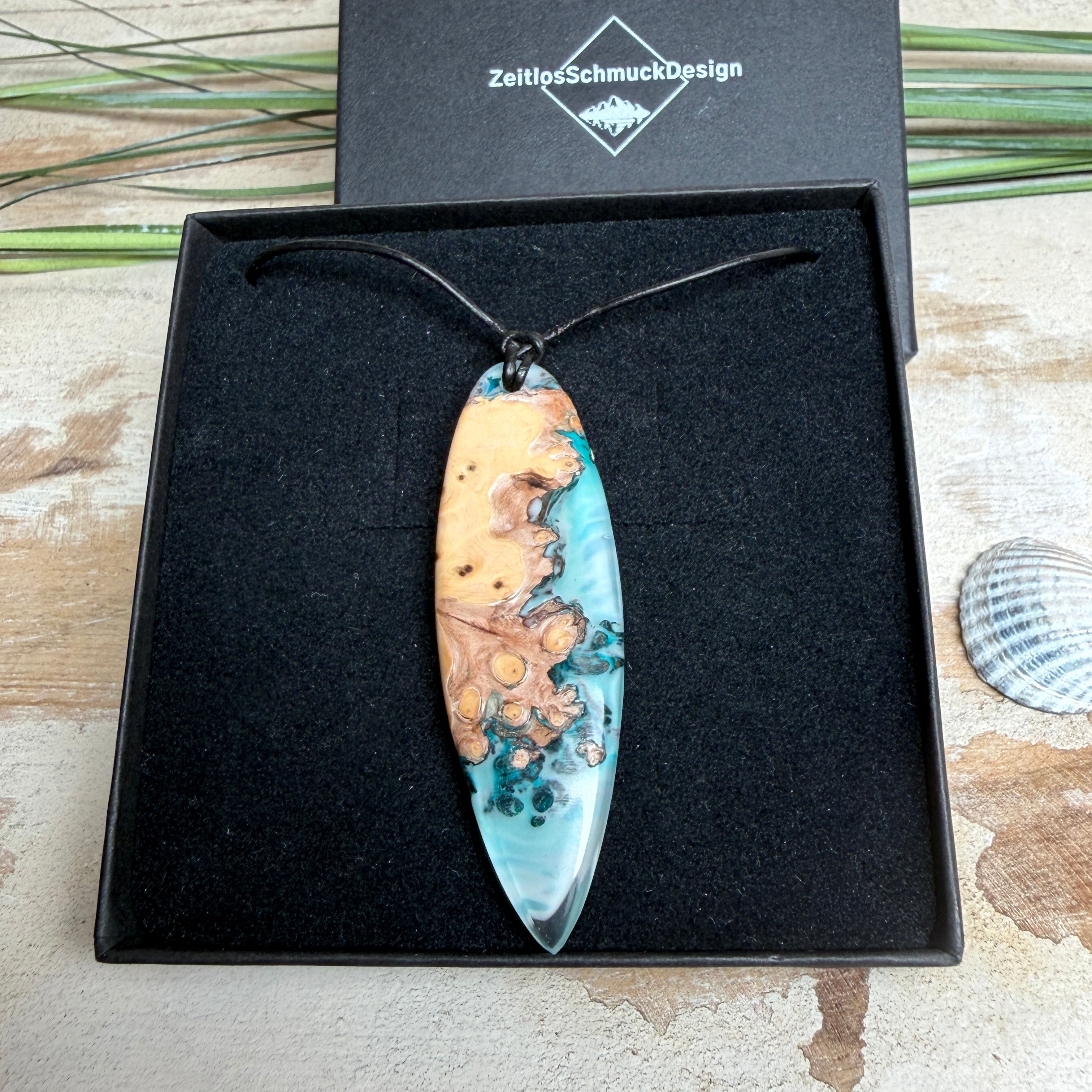 Handgefertigter, länglicher Surfbrett-Anhänger aus naturbelassenem Maserholz und transparentem Harz in Lagunen-Türkis, an schwarzem Lederband mit Knoten – maritimer Unikatschmuck für Damen und Herren, dekorativ mit Stein und Muschel inszeniert.