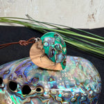 Herzförmiger Anhänger aus Olivenholz mit aquamarinfarbenem Harz und eingelegten Paua-Muschelstücken (Abalone), irisierender Blau-Grün-Schimmer. Der Anhänger hängt an einem braunen, verstellbaren Lederband mit Schiebeknoten; Aufhängung aus Edelstahl. Produktfoto vor maritimer Deko/Abalone-Schale.