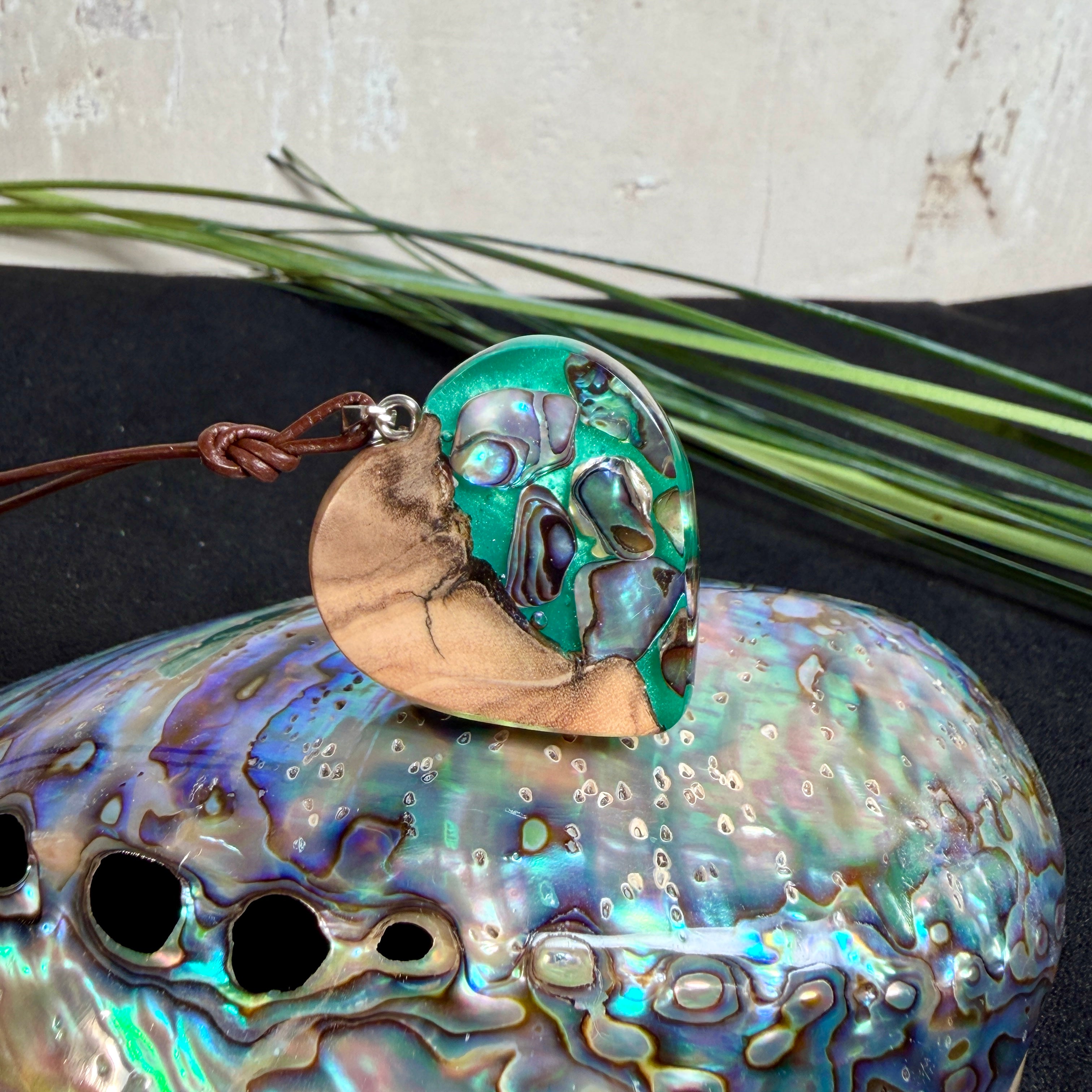 Herzförmiger Anhänger aus Olivenholz mit aquamarinfarbenem Harz und eingelegten Paua-Muschelstücken (Abalone), irisierender Blau-Grün-Schimmer. Der Anhänger hängt an einem braunen, verstellbaren Lederband mit Schiebeknoten; Aufhängung aus Edelstahl. Produktfoto vor maritimer Deko/Abalone-Schale.