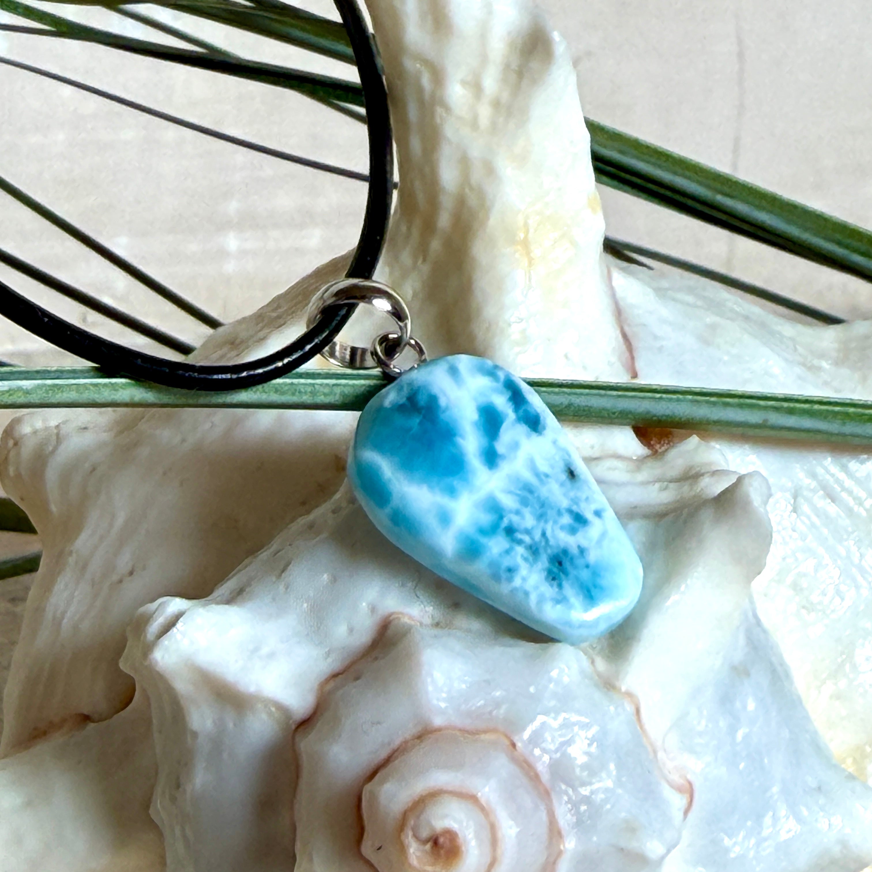 Larimar Freeform-Anhänger auf schwarzer Lederkette. Beidseitig tragbarer Trommelstein, ca. 2 x 1,8 cm groß, mit Silberöse. Der seltene, kostbare Larimar zeigt lebendige Blau- und Weißnuancen wie karibisches Lagunenwasser. Lederkette 45 cm mit Edelstahl-Karabiner und 5 cm Edelstahl-Verlängerung, fotografiert vor einer großen Muschel im maritimen Stil.