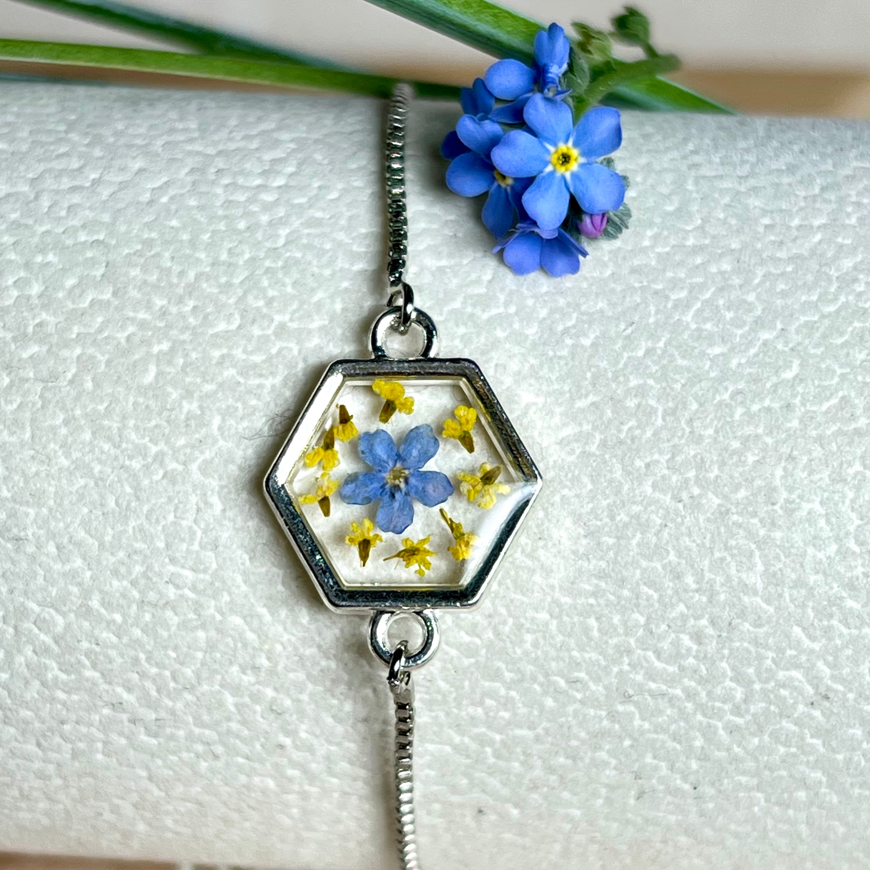 „Detailaufnahme eines handgefertigten Armbands mit echter blauer Vergissmeinnicht und feinen gelben Blüten in Harz, eingefasst in sechseckiger versilberter Lünette. Verstellbares Edelstahlarmband mit funkelnden Zirkonia-Endperlen – liebevolles Natur-Schmuckstück, ideal als Geschenk für Frauen.“