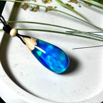 Handgefertigter Tropfenanhänger aus Maserholz und Harz mit synthetischem Opal. Die Farben schimmern je nach Lichteinfall in leuchtendem Blau und Grün – inspiriert von Polarlichtern. Der Anhänger ist ca. 3,9 × 1,8 cm groß und hängt an einem verstellbaren Lederband mit Holzperlen. Jedes Stück ist ein Unikat mit einzigartiger Holzmaserung. Ideal als Geschenk oder stilvoller Begleiter im Alltag.