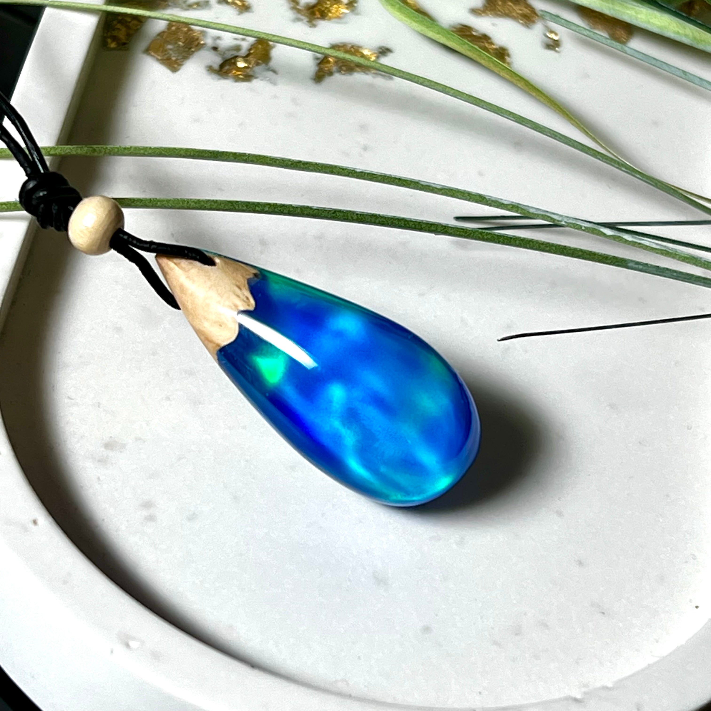 Handgefertigter Tropfenanhänger aus Maserholz und Harz mit synthetischem Opal. Die Farben schimmern je nach Lichteinfall in leuchtendem Blau und Grün – inspiriert von Polarlichtern. Der Anhänger ist ca. 3,9 × 1,8 cm groß und hängt an einem verstellbaren Lederband mit Holzperlen. Jedes Stück ist ein Unikat mit einzigartiger Holzmaserung. Ideal als Geschenk oder stilvoller Begleiter im Alltag.