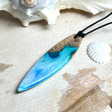 Surfbrett-Anhänger handgefertigt aus Maserholz & Harz in Blau-Weiß. Größe ca. 6,3 × 2 cm. Lederband verstellbar 26–47 cm, Schiebeknoten mit Holzperlen.