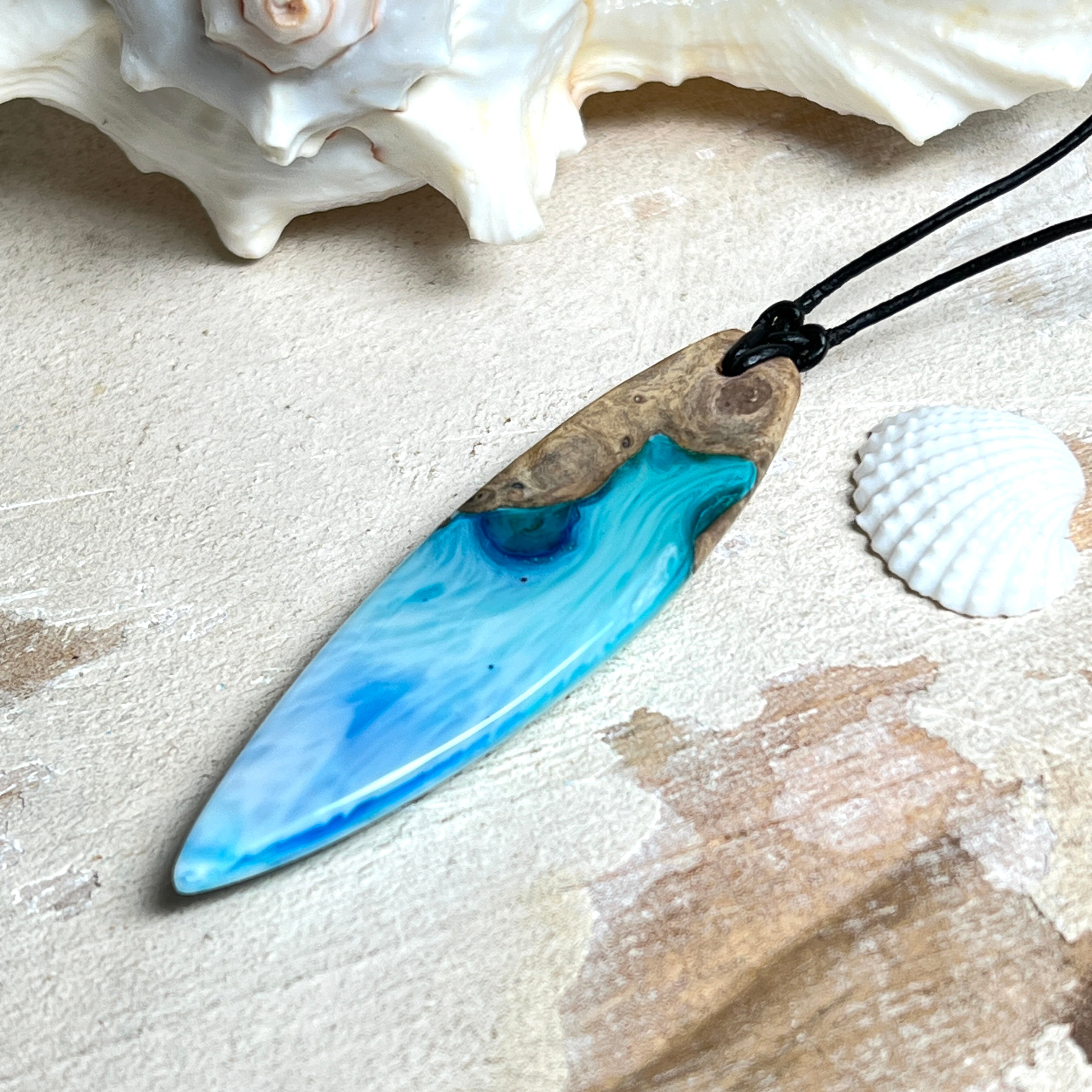 Surfbrett-Anhänger handgefertigt aus Maserholz & Harz in Blau-Weiß. Größe ca. 6,3 × 2 cm. Lederband verstellbar 26–47 cm, Schiebeknoten mit Holzperlen.