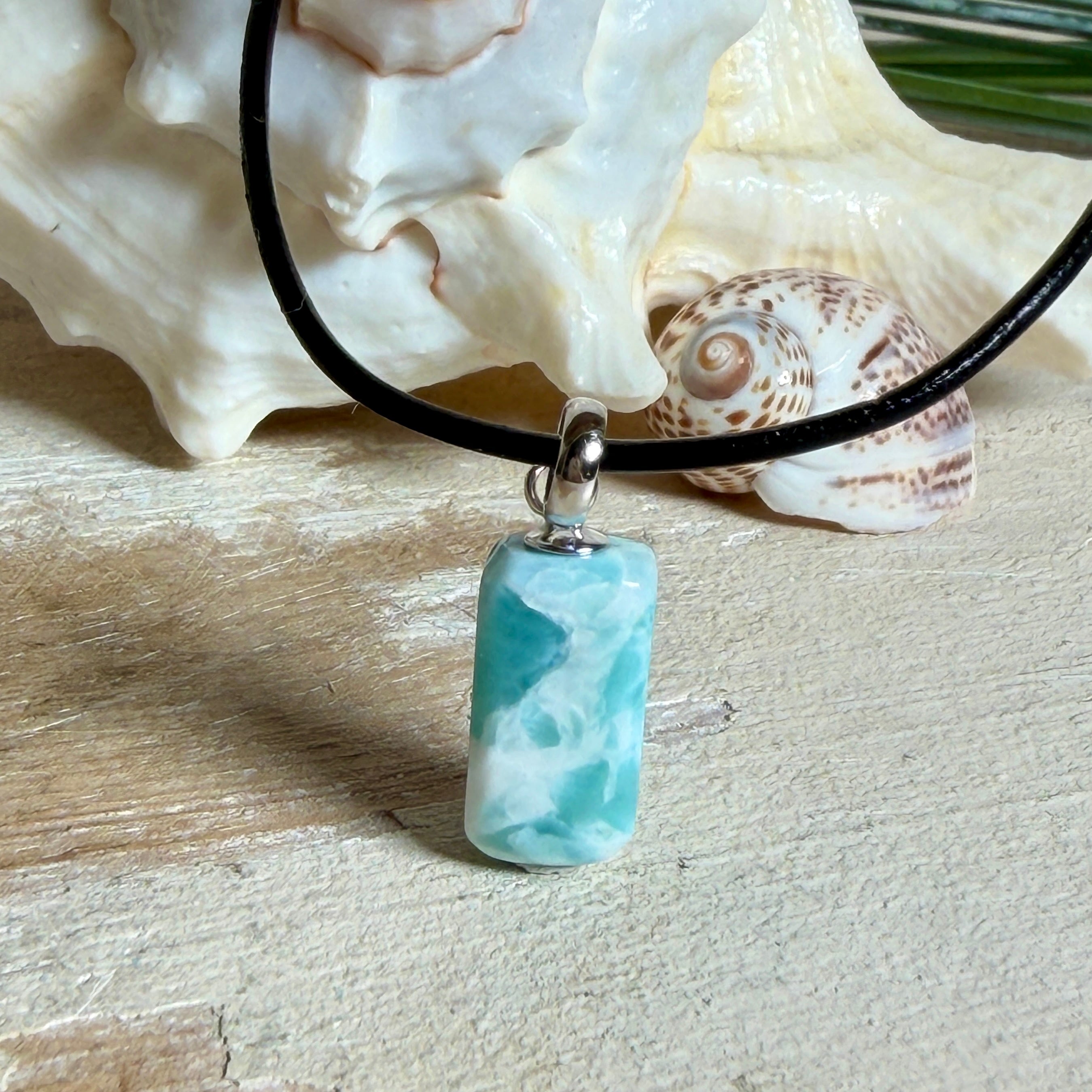 Larimar Freeform Anhänger an schwarzer Lederkette. Natürlicher, rechteckiger Larimar-Stein (ca. 1,5 × 1 cm) in Türkis-Blau mit weißen Strukturen, eingefasst in eine rhodinierte Silberöse. Beidseitig tragbar, an 45 cm Lederkette mit Edelstahl-Karabinerverschluss und 5 cm Verlängerung. Maritimes, zurückhaltend elegantes Schmuckstück mit der Optik karibischen Meerwassers.