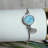 Verstellbares Edelstahlarmband mit Zirkoniasteinchen und Schiebeverschluss und rundem Anhänger (ca. 2,5 × 1,7 cm) mit echten Larimar-Edelstein-Splittern in Harz. Zusätzlich ein Charm „Blume des Lebens“ aus 925 Silber. Handgefertigtes Unikat in Türkis- und Meeresblautönen – Schmuckstück in Nahaufnahme bzw. als Tragebild am Handgelenk.