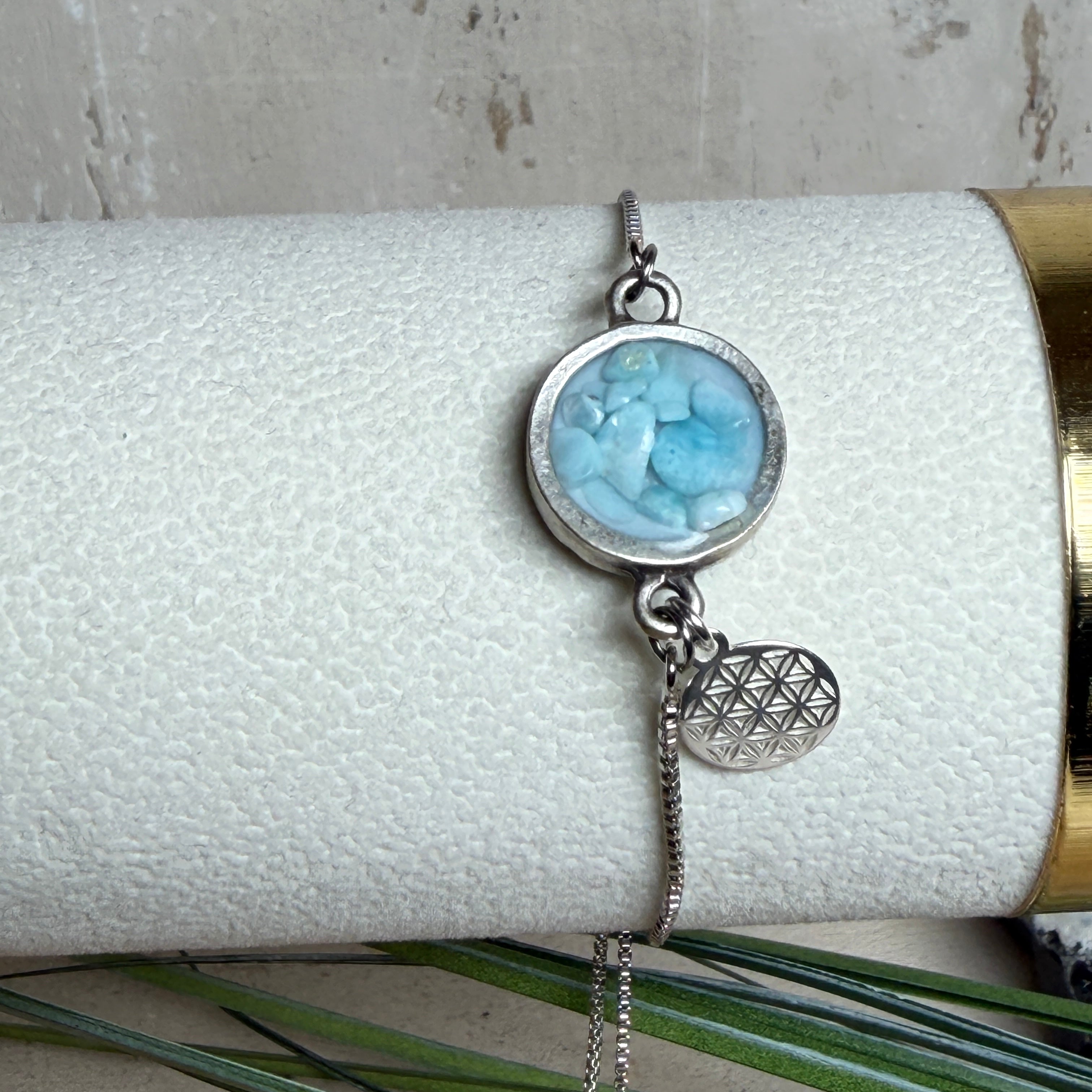 Verstellbares Edelstahlarmband mit Zirkoniasteinchen und Schiebeverschluss und rundem Anhänger (ca. 2,5 × 1,7 cm) mit echten Larimar-Edelstein-Splittern in Harz. Zusätzlich ein Charm „Blume des Lebens“ aus 925 Silber. Handgefertigtes Unikat in Türkis- und Meeresblautönen – Schmuckstück in Nahaufnahme bzw. als Tragebild am Handgelenk.