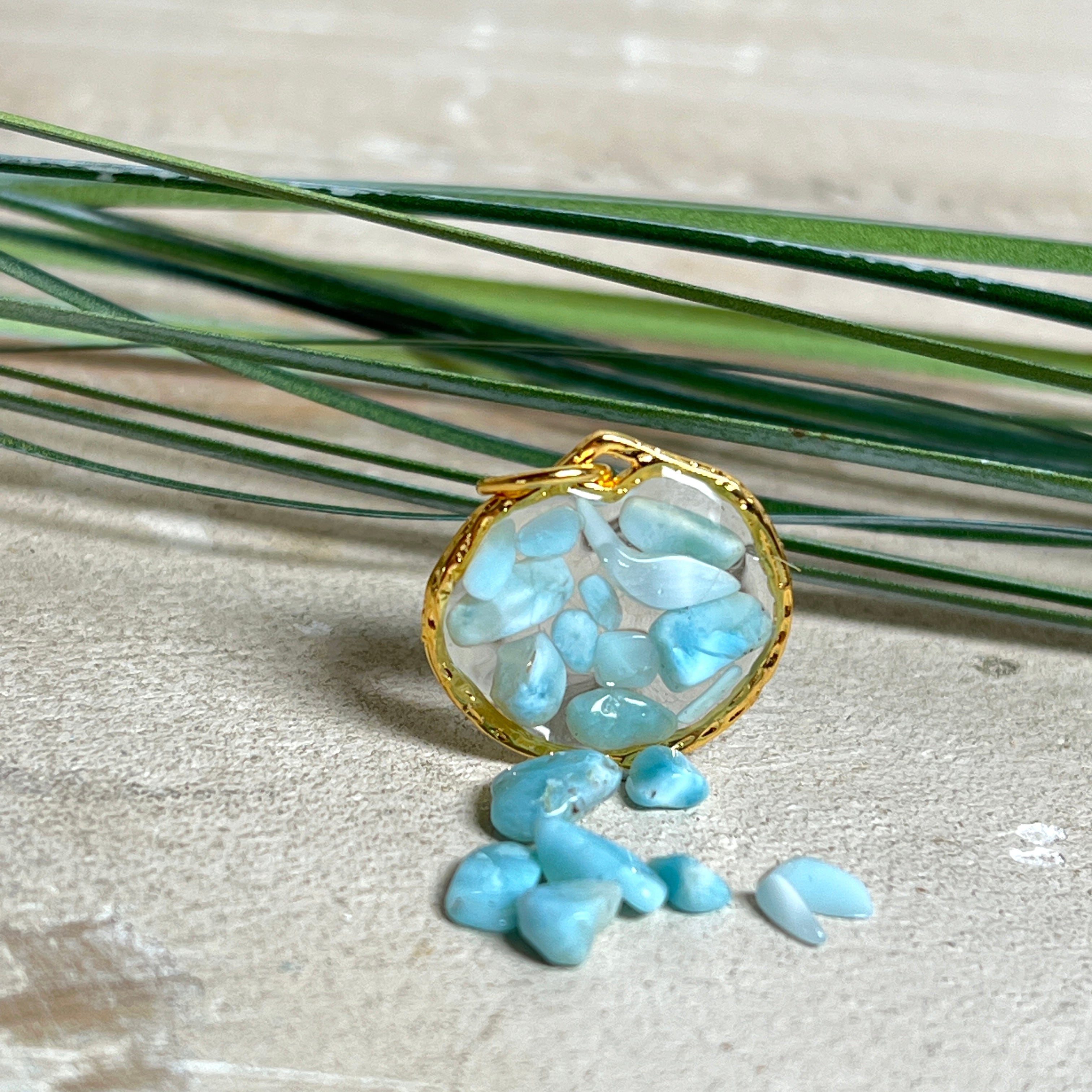 Runder Anhänger „Golden Atlantis Dream“ mit kleinen Larimar-Edelsteinchen in klarem Resin, gefasst in einer 24K vergoldeten Zinklegierung in Shiny Gold. Die Larimarsteine zeigen sanfte Blau- und Weißtöne wie karibische Wellen. Durchmesser ca. 22 mm, handgefertigtes Unikat, fotografiert mit Larimarsteinchen, Muscheln und einer 2-Euro-Münze zur Größenorientierung auf maritimem Untergrund.