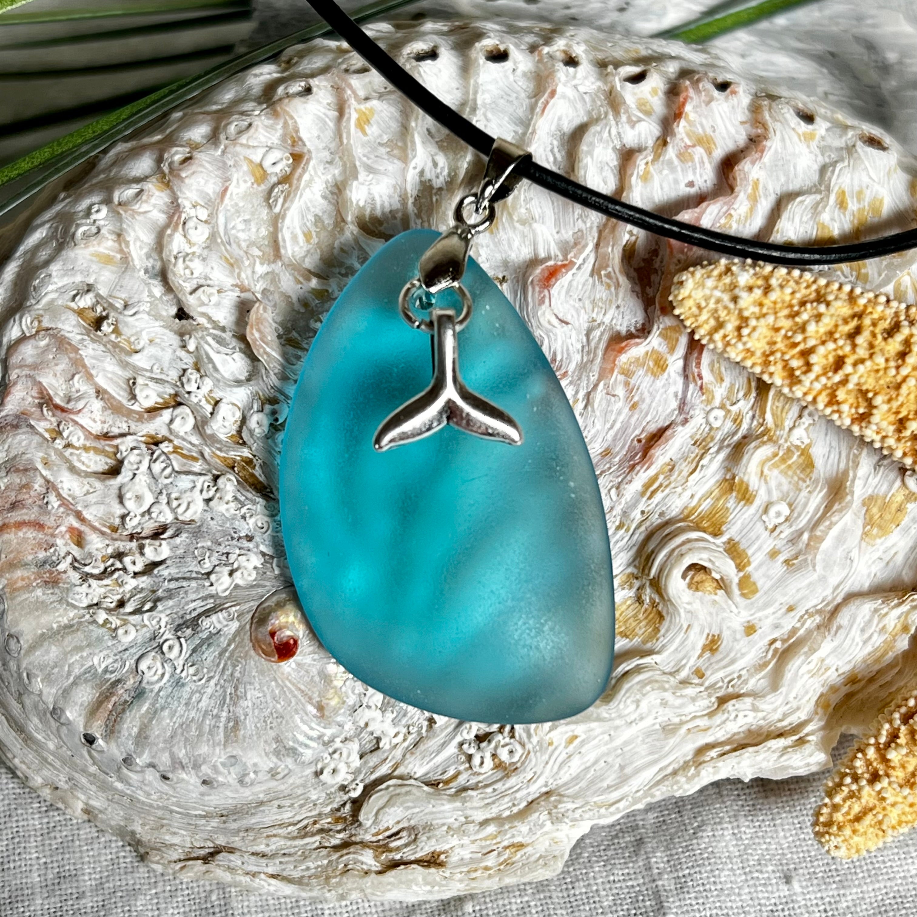 Edle Halskette „Blue Essence“ mit handgefertigtem türkisfarbenem Seeglas-Anhänger und Edelstahl-Flosse. Die Kette besteht aus schwarzer Lederkordel mit Karabinerverschluss und Verlängerung aus Edelstahl. Das matte Seeglas reflektiert Licht wie Meereswasser – ideal für Strandliebhaber und Naturliebhaber.