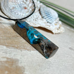 Rechteckiger Stabanhänger aus Maserholz und glasklarem Epoxidharz in tiefen Blau- und Türkistönen mit organischer Lagunen/Riff-Optik. Hochglanzpoliert, ca. 4,6 × 1,2 cm. Der Anhänger hängt an einer schwarzen Lederkette mit Edelstahl-Karabiner und 5-cm-Verlängerung (45 + 5 cm); die Anhängerschlaufe ist ebenfalls aus Edelstahl. Handgefertigtes Unikat, Lieferung in edler Schmuckbox – maritimer Look.