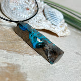 Rechteckiger Stabanhänger aus Maserholz und glasklarem Epoxidharz in tiefen Blau- und Türkistönen mit organischer Lagunen/Riff-Optik. Hochglanzpoliert, ca. 4,6 × 1,2 cm. Der Anhänger hängt an einer schwarzen Lederkette mit Edelstahl-Karabiner und 5-cm-Verlängerung (45 + 5 cm); die Anhängerschlaufe ist ebenfalls aus Edelstahl. Handgefertigtes Unikat, Lieferung in edler Schmuckbox – maritimer Look.