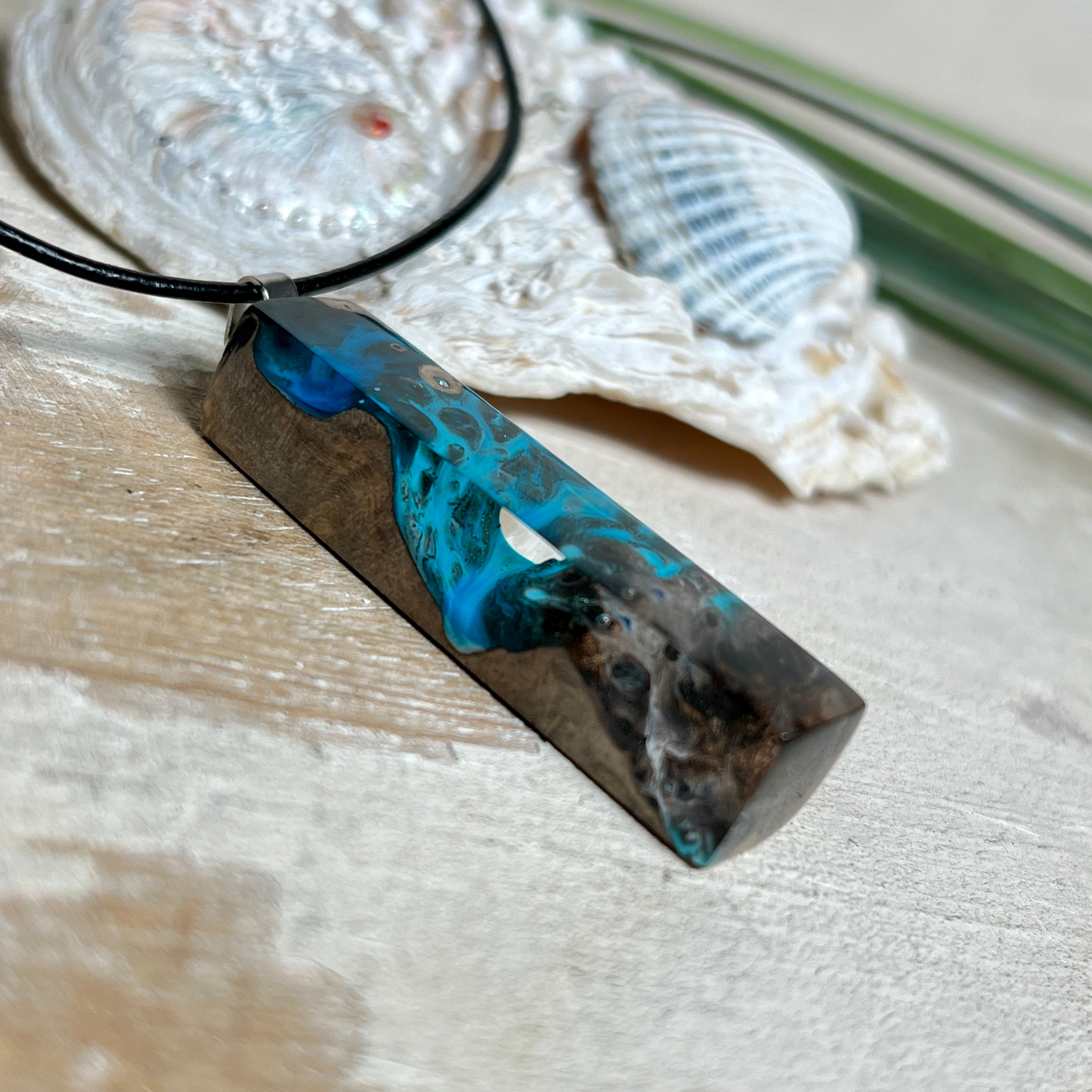 Rechteckiger Stabanhänger aus Maserholz und glasklarem Epoxidharz in tiefen Blau- und Türkistönen mit organischer Lagunen/Riff-Optik. Hochglanzpoliert, ca. 4,6 × 1,2 cm. Der Anhänger hängt an einer schwarzen Lederkette mit Edelstahl-Karabiner und 5-cm-Verlängerung (45 + 5 cm); die Anhängerschlaufe ist ebenfalls aus Edelstahl. Handgefertigtes Unikat, Lieferung in edler Schmuckbox – maritimer Look.