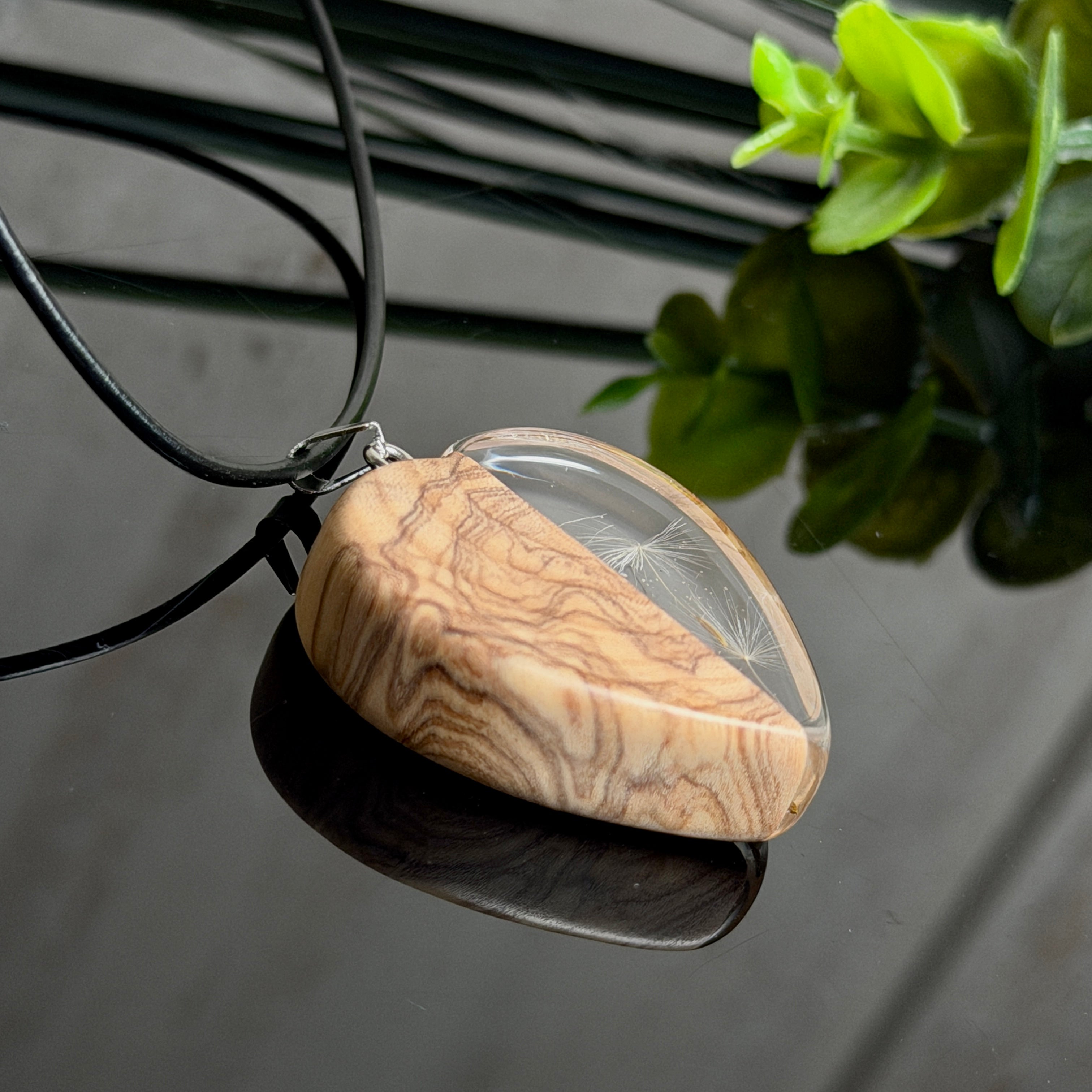 Pusteblumen Herzanhänger aus Olivenholz und glasklarem Epoxidharz, echte Löwenzahnsamen im Harz schwebend, schwarze Lederkette, handgefertigter Naturschmuck mit Spiegelung, dunkler Hintergrund