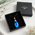 Aurora-Drop-Anhänger aus Maserholz und blau-grün schimmerndem Epoxidharz liegt in einer geöffneten schwarzen Schmuckbox von ZeitlosSchmuckDesign, daneben der Deckel mit Logo auf hellem Untergrund.