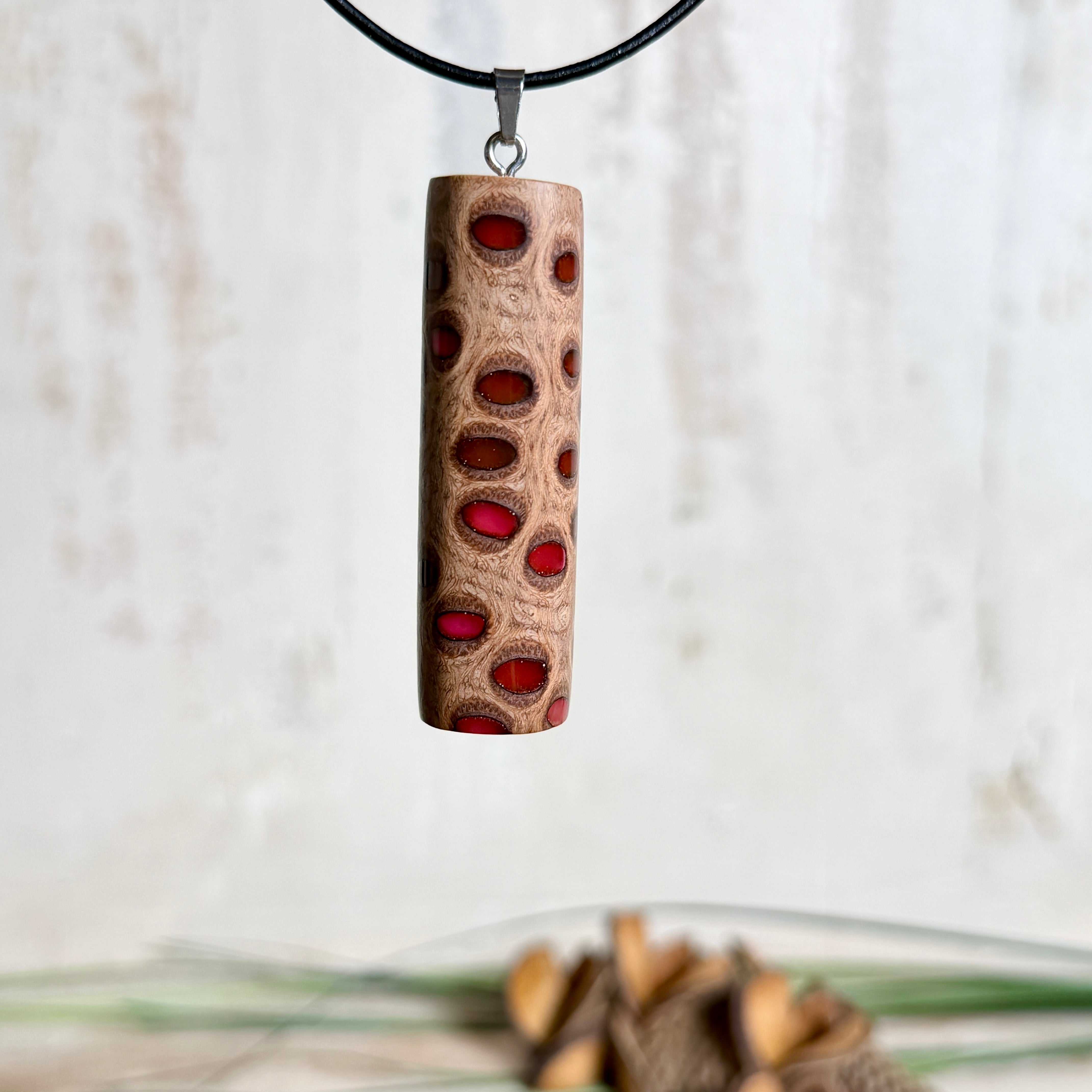 Handgefertigter Halsanhänger aus echtem Banksiazapfen, veredelt mit rotem Harz in den natürlichen Kammern; ca. 5,5 × 1,6 cm. Mit Anhängerschlaufe aus Edelstahl an schwarzer Lederkette (45 cm + 5 cm Verlängerung) mit Edelstahl-Karabiner. Detailaufnahme bzw. Tragebild vor neutralem, natürlichem Hintergrund.