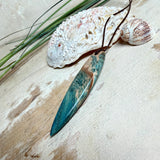 Unikat Anhänger aus stabilisiertem Holz mit türkisblauem Harzverlauf, schlanke Form, poliert. Verstellbares braunes Lederband 22–46 cm mit Schiebeknoten. Anhänger ca. 6,6 × 2 cm. Tragefoto am Hals und Produktfotos mit maritimer Deko