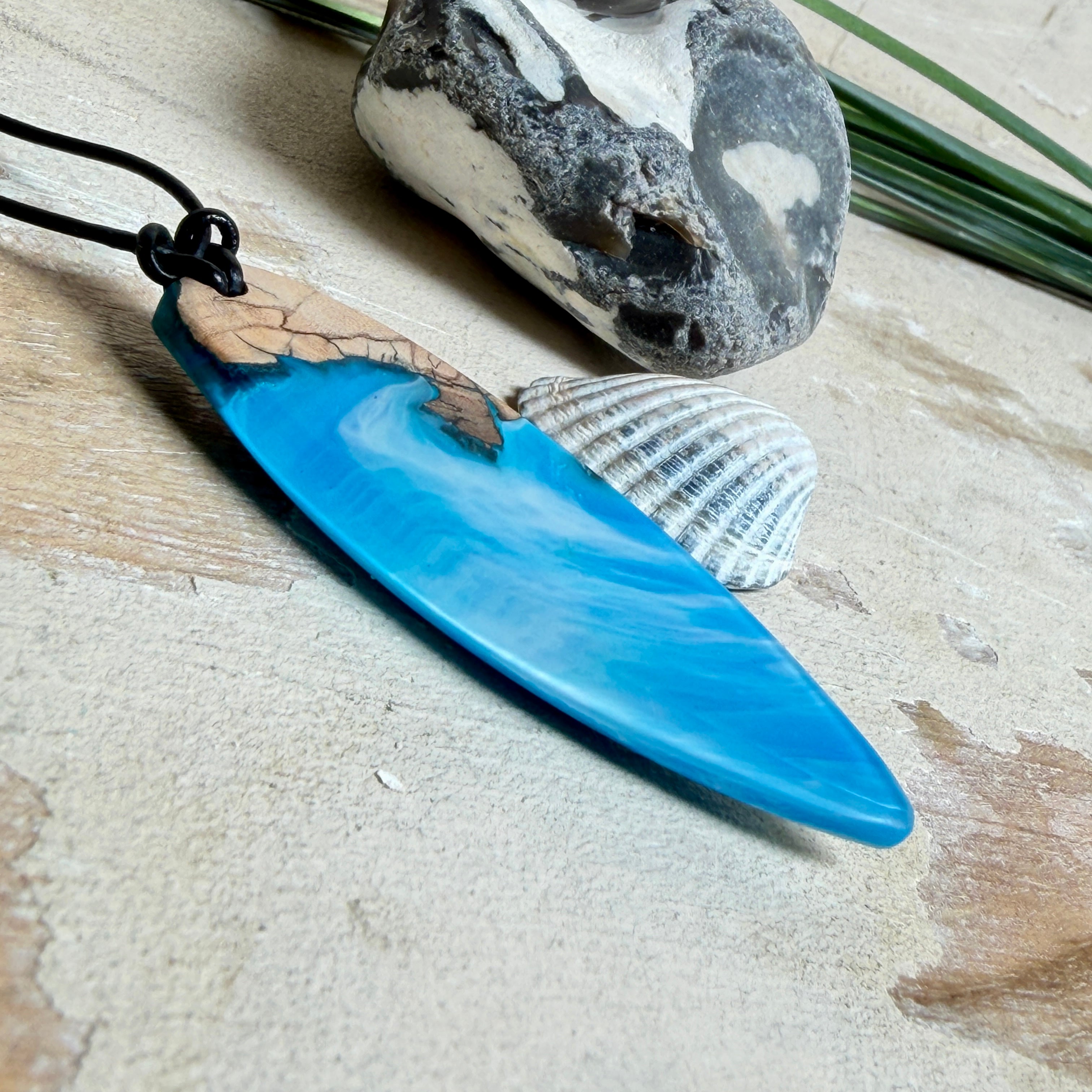 Handgefertigter Surfbrett-Anhänger aus warm gemasertem Olivenholz und Epoxidharz in Blau und Weiß, das an Meereswellen erinnert. Längliche, vorne polierte Form, ca. 6,2 × 2 cm, an verstellbarem schwarzem Lederband mit Knoten. Maritimer Unikatschmuck für Damen und Herren, dekorativ mit Stein und Muschel arrangiert.
