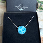 Feine Kette mit rundem Larimar-Cabochon in AA-Qualität. Der ca. 20 mm große Stein zeigt ein intensives Blau mit hellen, netzartigen Zeichnungen und ist mittig mit einer 2,5 mm Bohrung versehen, durch die eine zarte 925 Silberkette (45 cm, ca. 1 mm stark) geführt wird. 
