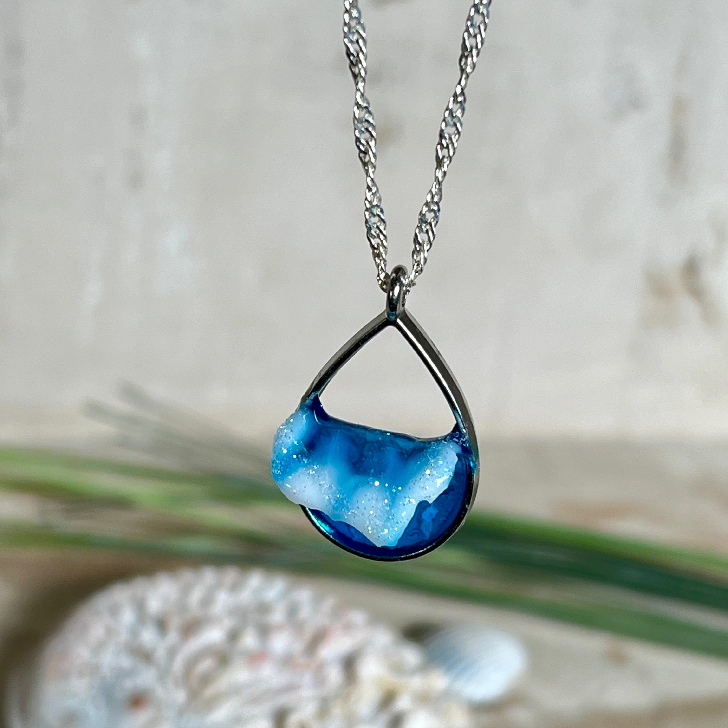 „Lagunenlicht“ – Tropfenanhänger mit handmodellierter 3D-Welle und feinem Glitzereffekt. Klarer Harzlook in Blau/Türkis, gefasst in Silber-Legierung. Größe ca. 3,5 × 2 cm. Geliefert an einer versilberten Kette (ca. 56 cm). Hochglänzend , maritimer Unikatcharakter.