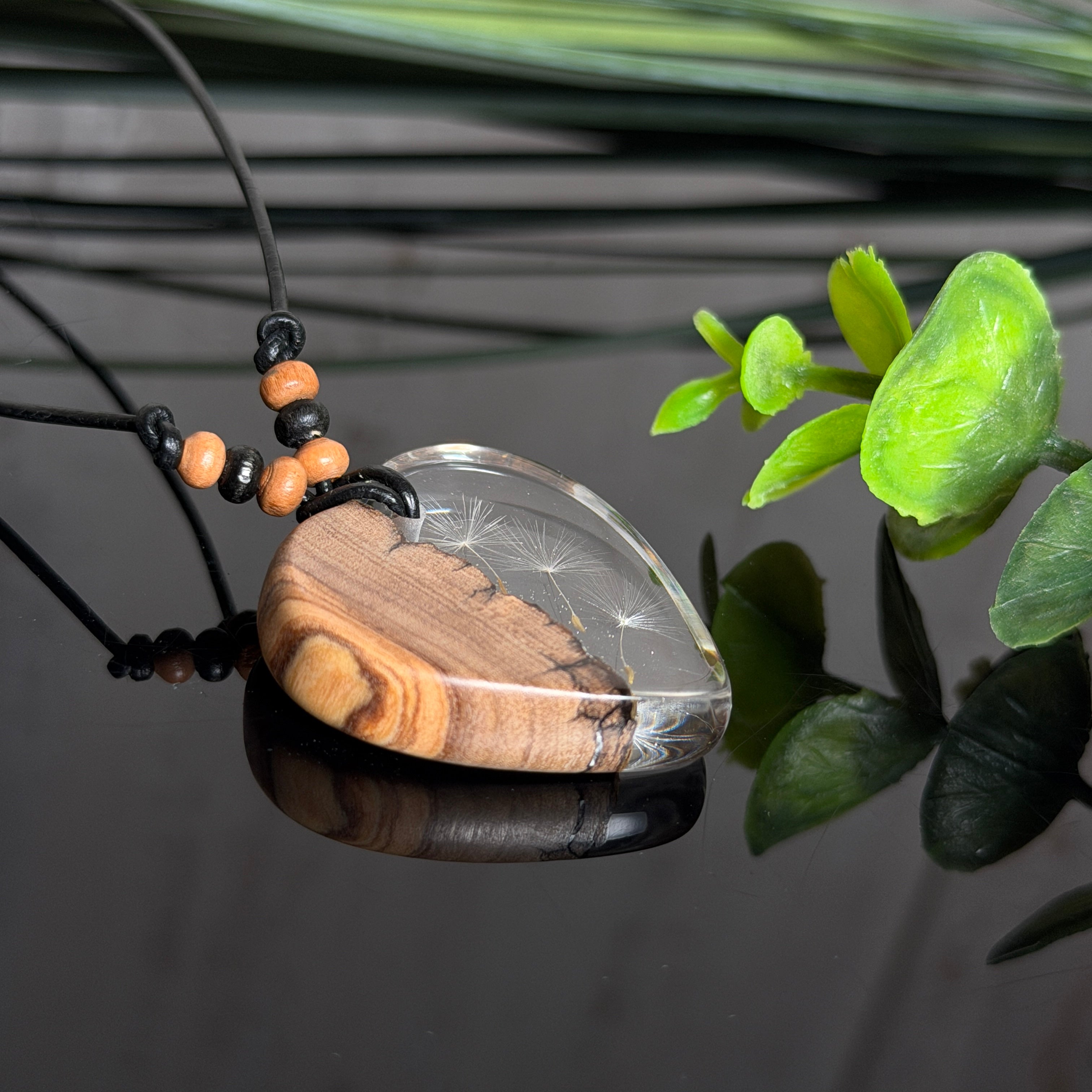 Handgefertigter Herzanhänger aus warm gemasertem Olivenholz und kristallklarem Epoxidharz mit schwebenden Pusteblumensamen. Schwarzes, verstellbares Lederband mit Schiebeknoten und naturfarbenen Holzperlen, Anhänger ca. 3,5 × 3,3 cm. Naturnaher Unikatschmuck von ZeitlosSchmuckDesign.