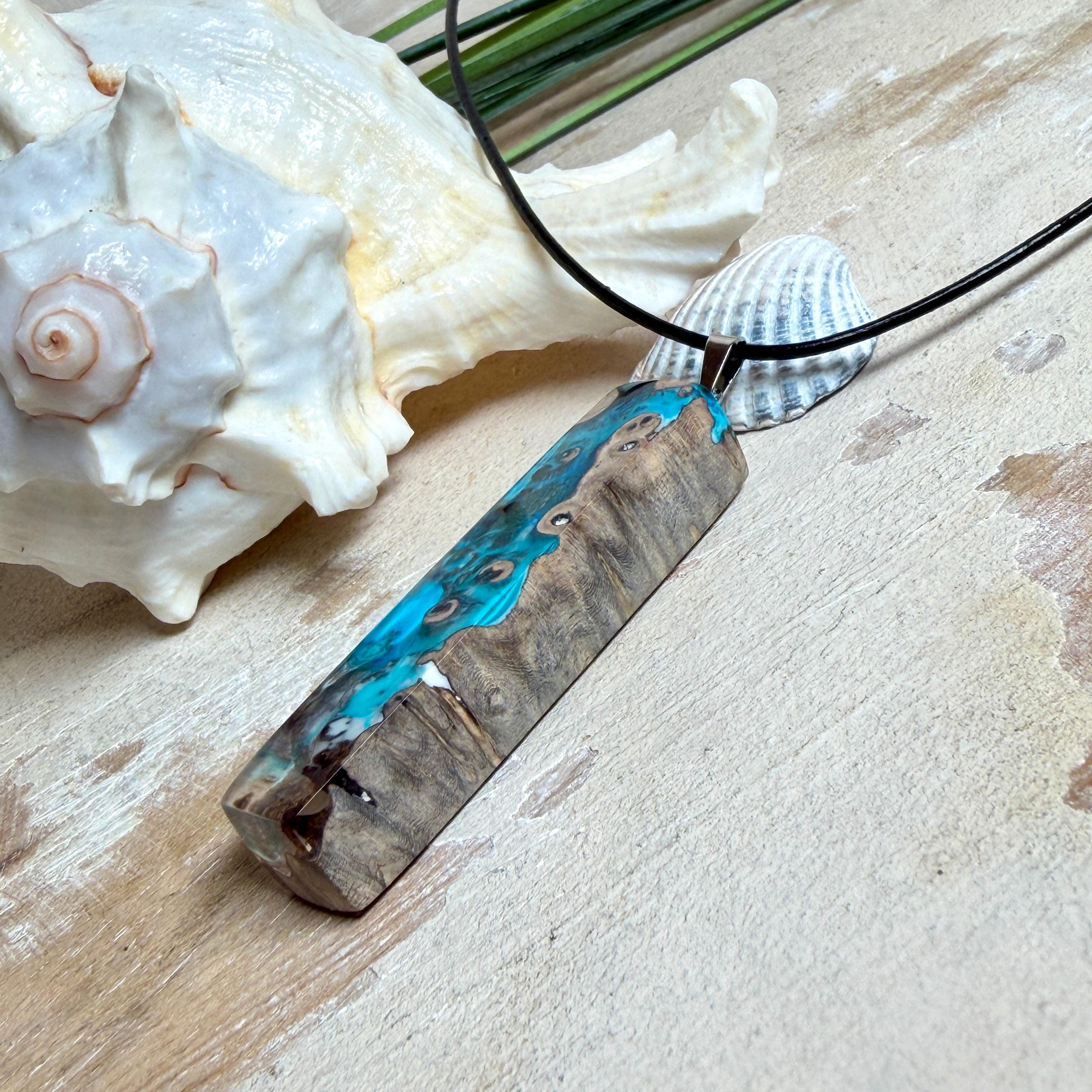 Rechteckiger Stabanhänger aus Maserholz und glasklarem Epoxidharz in tiefen Blau- und Türkistönen mit organischer Lagunen/Riff-Optik. Hochglanzpoliert, ca. 5,3 × 1,2 cm. Der Anhänger hängt an einer schwarzen Lederkette mit Edelstahl-Karabiner und 5-cm-Verlängerung (45 + 5 cm); die Anhängerschlaufe ist ebenfalls aus Edelstahl. Handgefertigtes Unikat, Lieferung in edler Schmuckbox – maritimer Look.