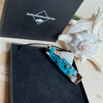 Rechteckiger Stabanhänger aus Maserholz und glasklarem Epoxidharz in tiefen Blau- und Türkistönen mit organischer Lagunen/Riff-Optik. Hochglanzpoliert, ca. 5,3 × 1,2 cm. Der Anhänger hängt an einer schwarzen Lederkette mit Edelstahl-Karabiner und 5-cm-Verlängerung (45 + 5 cm); die Anhängerschlaufe ist ebenfalls aus Edelstahl. Handgefertigtes Unikat, Lieferung in edler Schmuckbox – maritimer Look.