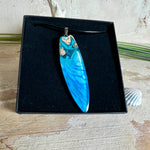 Surfbrett-Anhänger aus Maserholz und blau-weißem Epoxidharz, hochglanzpoliert, ca. 6,5 × 2 cm. An schwarzer Lederkette mit Edelstahl-Karabiner und 5-cm-Verlängerung; Edelstahl-Öse. Im Tageslicht lagunenblau, unter Schwarzlicht fluoreszierend – ideal als Party-/Festival-Schmuck. Handgefertigtes Unikat, Lieferung in edler Schmuckbox.