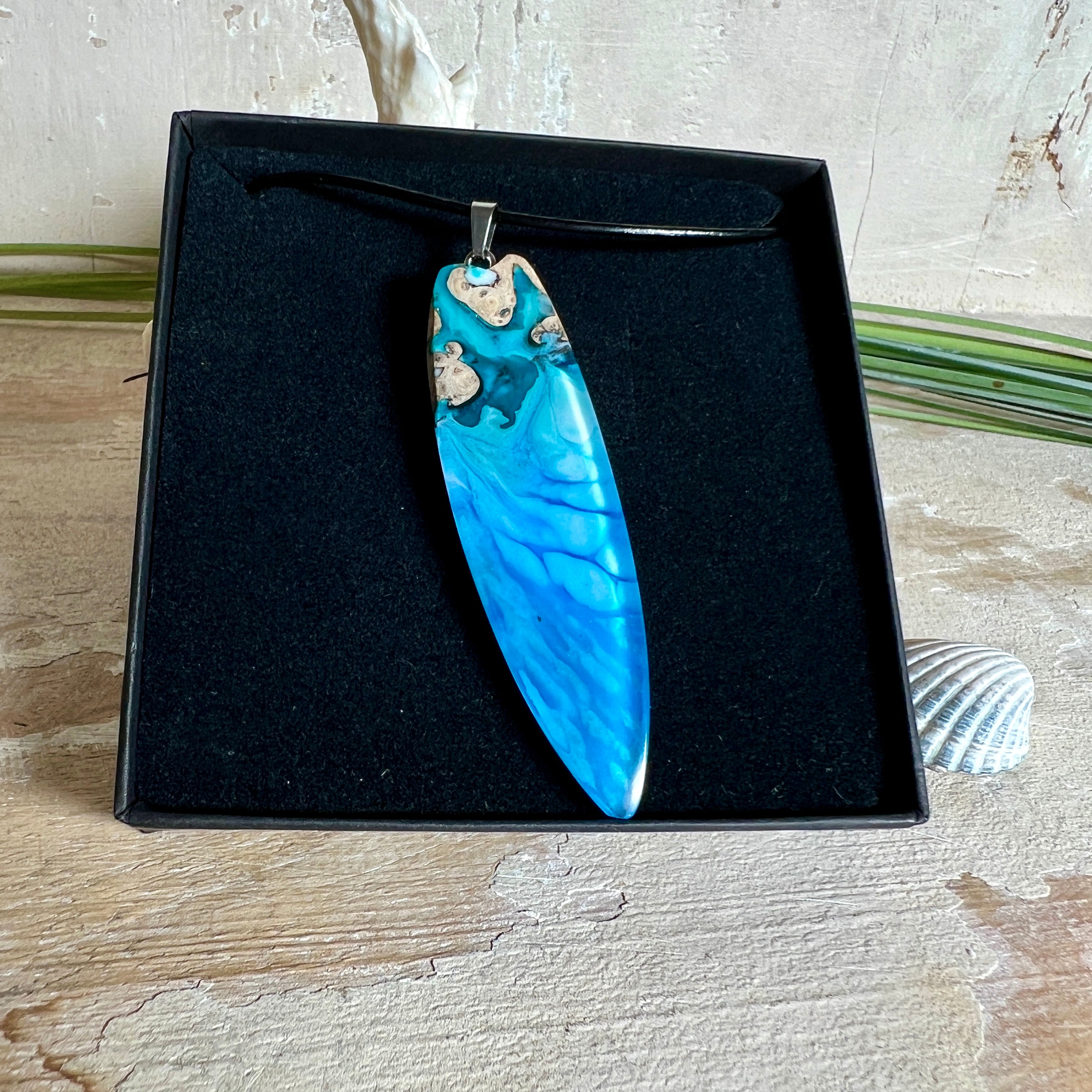 Surfbrett-Anhänger aus Maserholz und blau-weißem Epoxidharz, hochglanzpoliert, ca. 6,5 × 2 cm. An schwarzer Lederkette mit Edelstahl-Karabiner und 5-cm-Verlängerung; Edelstahl-Öse. Im Tageslicht lagunenblau, unter Schwarzlicht fluoreszierend – ideal als Party-/Festival-Schmuck. Handgefertigtes Unikat, Lieferung in edler Schmuckbox.
