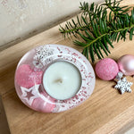 Rosa Teelichthalter aus klarem Epoxidharz, ca. 8 × 6,5 cm, Höhe ca. 4 cm. Im Harz sind kleine rosa Weihnachtskugeln, ein Stern, eine Schneeflocke und rosa Granulat eingegossen. Ovaler Halter für ein Teelicht, Unterseite mit vier runden Korkpads als Möbelschoner. Fotografiert auf Holz mit Tannenzweig und rosa Kugeln als weihnachtlicher Dekohintergrund.