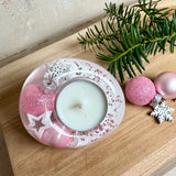 Rosa Teelichthalter aus klarem Epoxidharz, ca. 8 × 6,5 cm, Höhe ca. 4 cm. Im Harz sind kleine rosa Weihnachtskugeln, ein Stern, eine Schneeflocke und rosa Granulat eingegossen. Ovaler Halter für ein Teelicht, Unterseite mit vier runden Korkpads als Möbelschoner. Fotografiert auf Holz mit Tannenzweig und rosa Kugeln als weihnachtlicher Dekohintergrund.