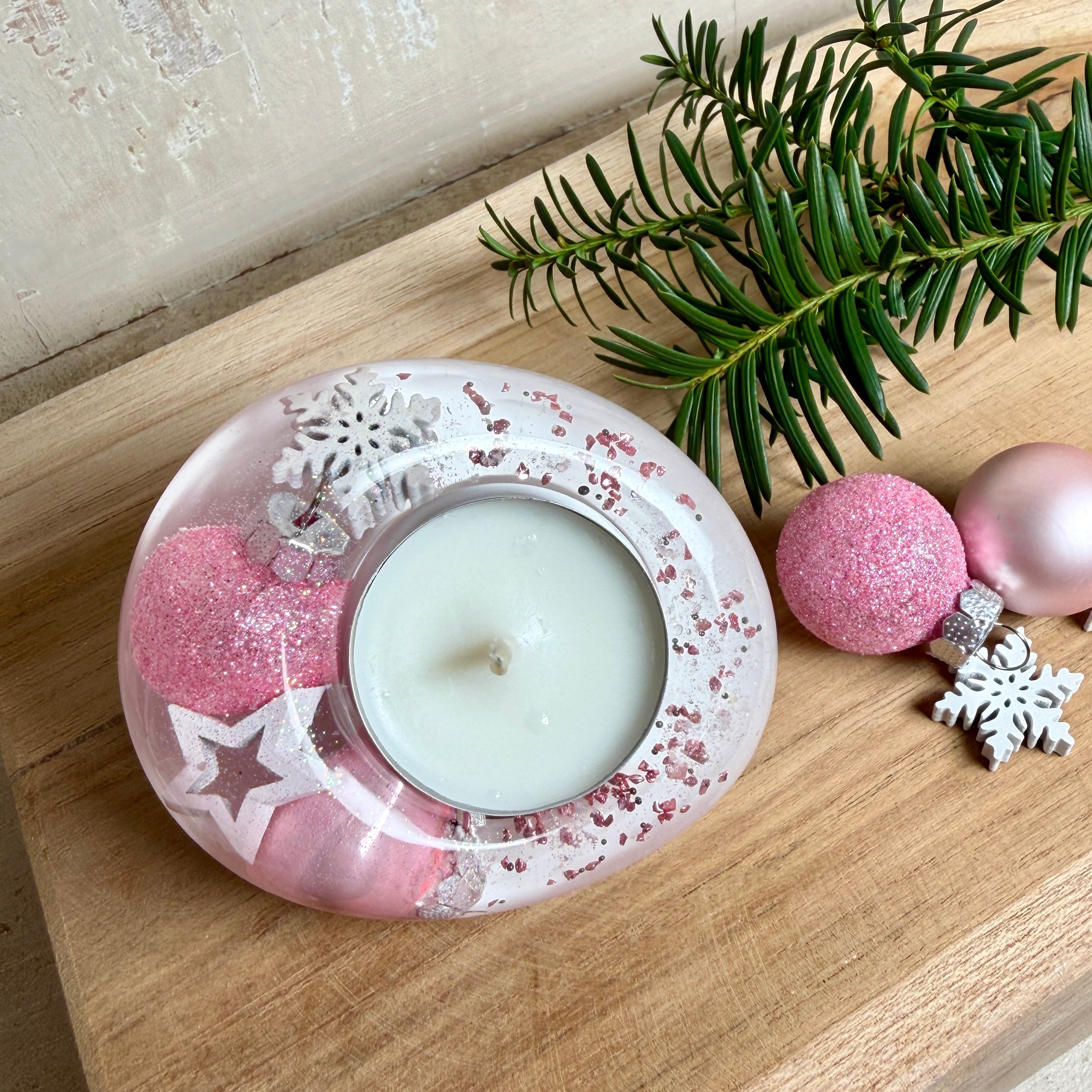 Rosa Teelichthalter aus klarem Epoxidharz, ca. 8 × 6,5 cm, Höhe ca. 4 cm. Im Harz sind kleine rosa Weihnachtskugeln, ein Stern, eine Schneeflocke und rosa Granulat eingegossen. Ovaler Halter für ein Teelicht, Unterseite mit vier runden Korkpads als Möbelschoner. Fotografiert auf Holz mit Tannenzweig und rosa Kugeln als weihnachtlicher Dekohintergrund.