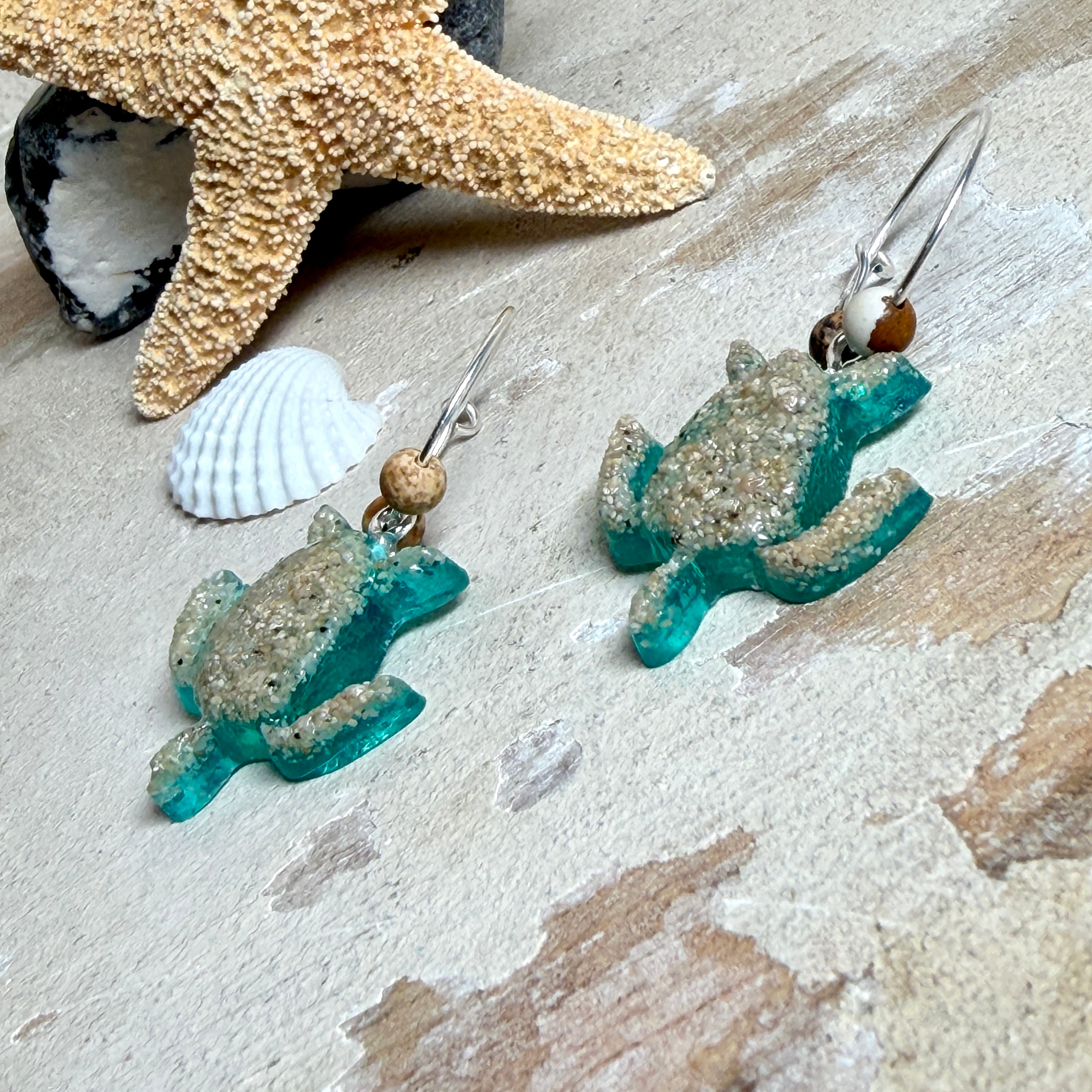 Türkisfarbene Schildkröten-Creolen an feinen 16-mm-Ringen aus 925 Silber, jeweils mit echtem Strandsand im Harz eingebettet und mit kleinen Jaspis-Edelsteinperlen verziert; handgefertigtes Unikat mit sommerlich-maritimem Look, fotografiert als Freisteller, Nahaufnahme oder auf dekorativem Untergrund.