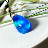 Handgefertigter Tropfenanhänger aus Maserholz und Harz mit synthetischem Opal. Die Farben schimmern je nach Lichteinfall in leuchtendem Blau und Grün – inspiriert von Polarlichtern. Der Anhänger ist ca. 3,9 × 1,8 cm groß und hängt an einem verstellbaren Lederband mit Holzperlen. Jedes Stück ist ein Unikat mit einzigartiger Holzmaserung. Ideal als Geschenk oder stilvoller Begleiter im Alltag.