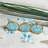 Runder Anhänger „Golden Atlantis Dream“ mit kleinen Larimar-Edelsteinchen in klarem Resin, gefasst in einer 24K vergoldeten Zinklegierung in Shiny Gold. Die Larimarsteine zeigen sanfte Blau- und Weißtöne wie karibische Wellen. Durchmesser ca. 22 mm, handgefertigtes Unikat, fotografiert mit Larimarsteinchen, Muscheln und einer 2-Euro-Münze zur Größenorientierung auf maritimem Untergrund.