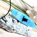 Heller Holzhintergrund: Surfbrett Anhänger aus hellblauem Epoxidharz mit schwarzer Schildkröten-Silhouette und weißem Welleneffekt, schwarze Lederkette, handgefertigter maritimer Naturschmuck.