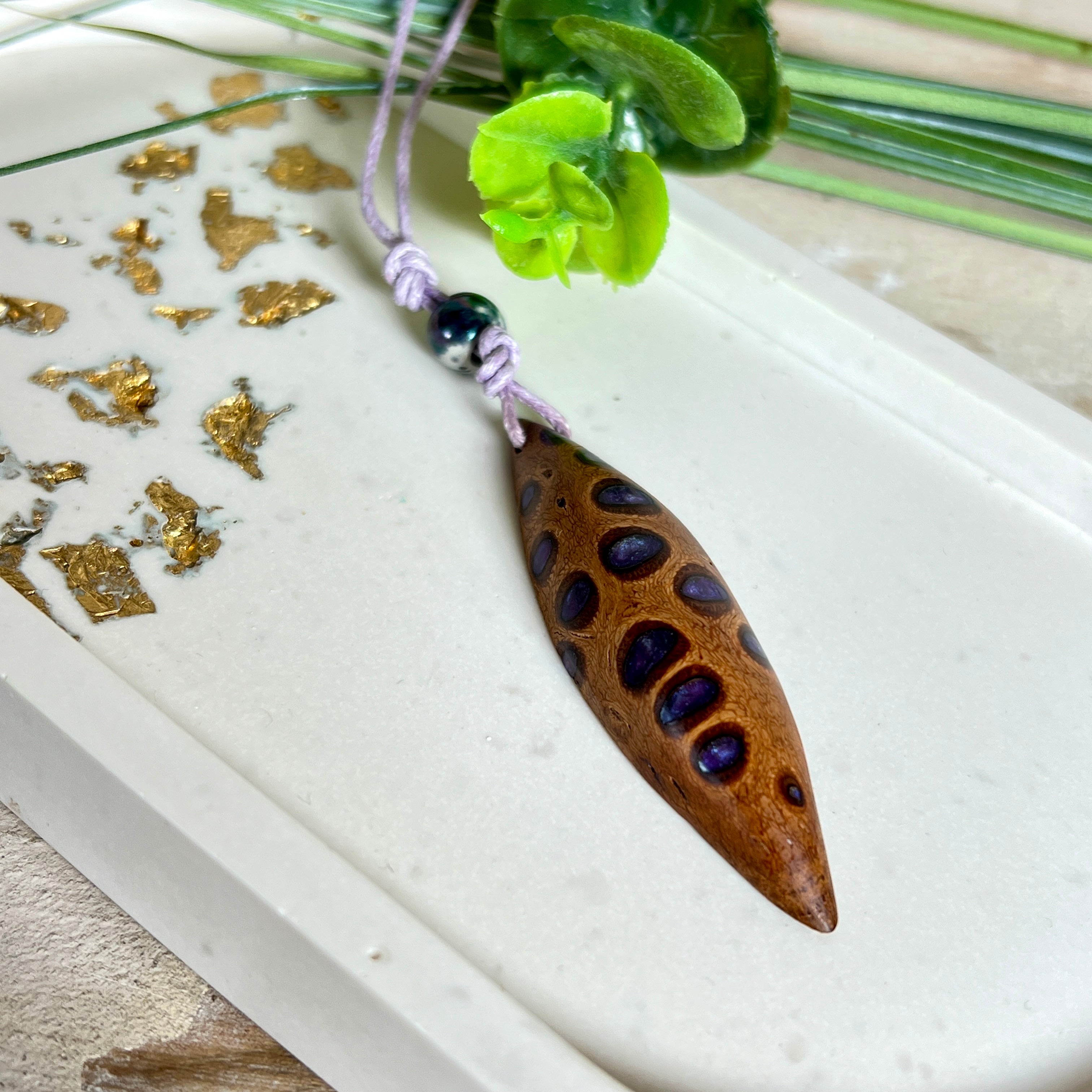Handgefertigter Anhänger aus einem australischen Banksiazapfen mit eingelassenem metallic-violettem Harz. Der ovale, tropfenförmige Anhänger ist ca. 5,5 cm lang und wird mit einem verstellbaren Baumwollband geliefert. Jedes Stück zeigt die einzigartige natürliche Struktur des Zapfens und schimmert in satten Lilatönen – ein stilvoller Schmuck mit organischem Charakter.