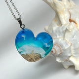 Ein herzförmiger Anhänger mit einer ganzen Ozeanwelt darin: Welle, Sand und Seestern in leuchtendem Blau und Türkis – handgegossen, einzigartig, unvergesslich. Mit versilberter Kette (46 cm) und Edelstahl-Karabiner.