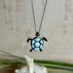 Feine Kette aus rhodiniertem 925 Silber mit Wasserschildkröten-Anhänger. Der ca. 25 x 18 mm große Anhänger zeigt eine Schildkröte, deren Panzer aus mehreren hellblauen Larimar-Inlays besteht. Seltener, kostbarer Larimar mit typischen wolkigen Strukturen, an einer ca. 45 cm langen, 1 mm starken, auf Platin rhodinierten Silberkette. Fotografiert in schwarzer Schmuckbox und vor maritimer Dekoration mit Muscheln.